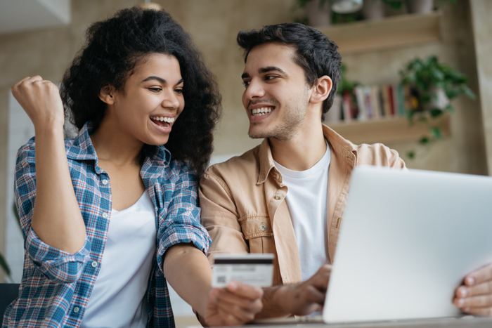 Happy couple shopping online, sitting at home. Beautiful African American woman holding credit card, attractive man booking tickets, ordering food on website