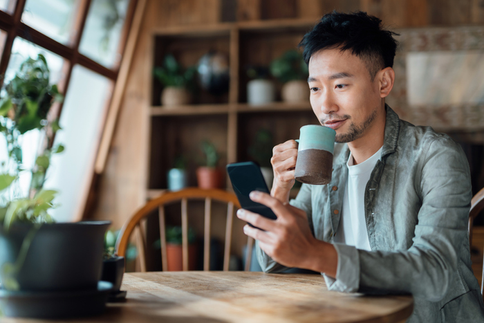 Young Asian man managing online banking with mobile app on smartphone, taking care of his money and finances while relaxing at home. Banking with technology